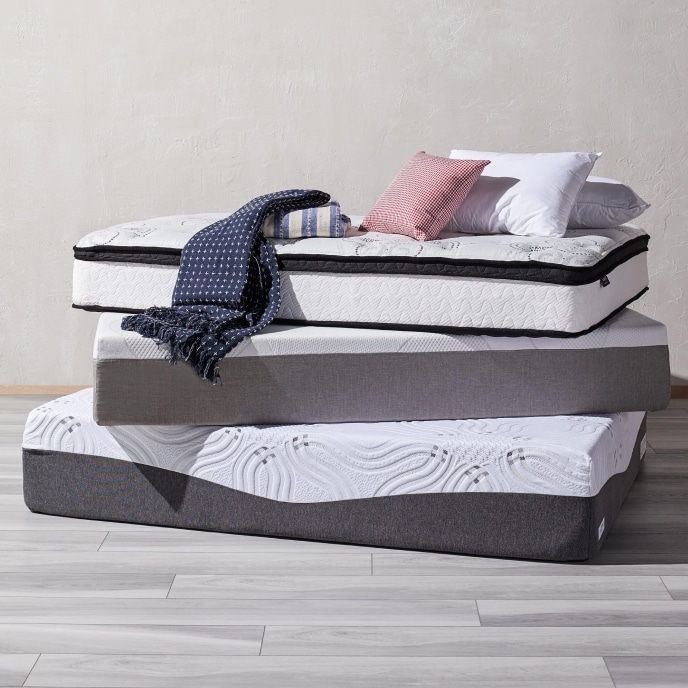 A stack of memory foam and pillowtop mattresses, topped with pillows and blue and white throw blankets.