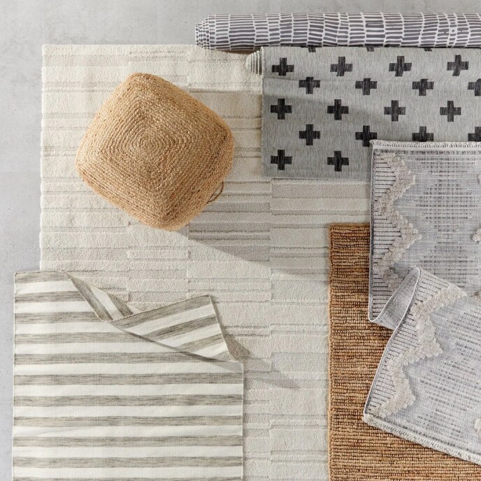 An overhead view of layered area rugs with geometric patterns, topped with a square, woven jute pouf ottoman.